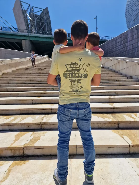 Padre de espaldas llevando en brazos a sus dos hijos pequeños al pie de unas escaleras, mirando hacia el futuro.